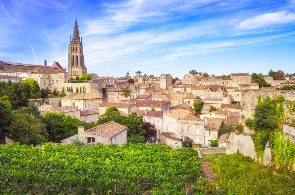 Vue sur Saint-Émilion : vignobles verdoyants, maisons en pierre et clocher historique sous un ciel bleu ensoleillé.