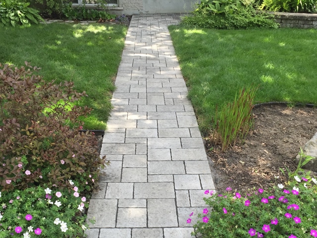 Allée de Jardin en Pavés : Aménagement Paysager Réussi Allée en pavés gris traversant un jardin fleuri avec pelouse verdoyante et massifs de fleurs roses et blanches.