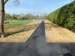 Aménagement d'une allée en goudron avec bordures blanches Allée en asphalte neuve avec bordures blanches traversant un parc entre une haie et un terrain de sport.