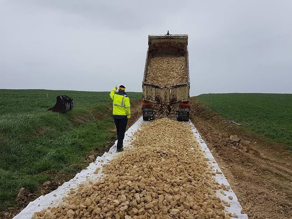 Construction de chemin : pose de géotextile et cailloux Un camion-benne décharge des cailloux sur un feutre géotextile pour créer un chemin rural entre deux champs.