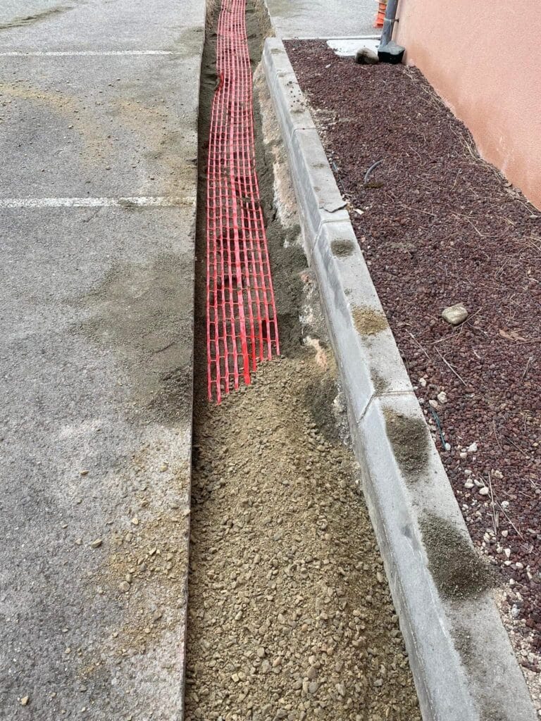 Pose de grillage avertisseur rouge en tranchée de parking Pose d'un grillage avertisseur rouge dans une tranchée de gravier pour la protection de réseaux électriques enterrés.