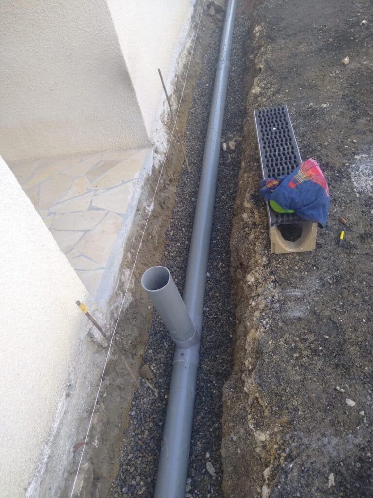 Installation Drainage Extérieur : Tuyaux PVC et Tranchée Installation d'un tuyau de drainage PVC gris dans une tranchée de gravier le long d'un mur de maison.