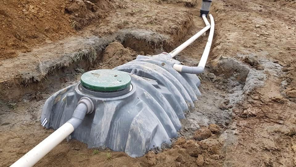 Installation fosse septique : pose d'un réservoir enterré Fosse septique en plastique gris avec couvercle vert et raccordements PVC dans une tranchée d'assainissement.