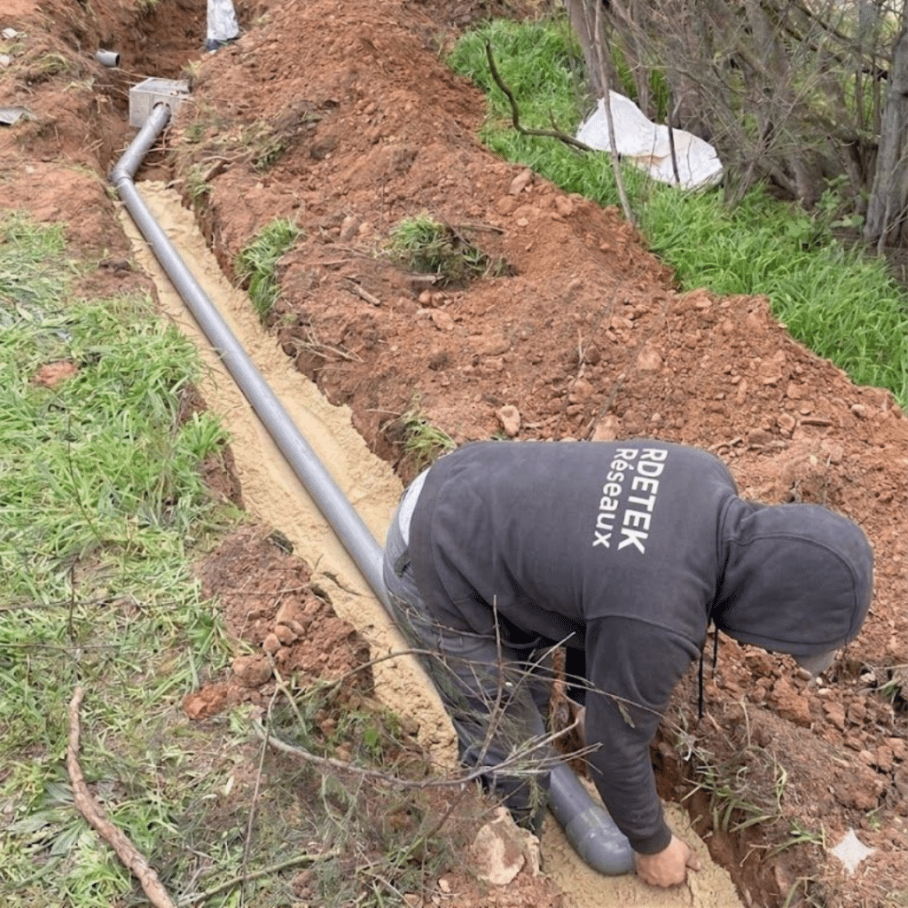 Installation de réseaux par RDETEK : pose de tuyau PVC Un professionnel de RDETEK Réseaux installe un tuyau d'évacuation gris dans une tranchée ensablée en extérieur.