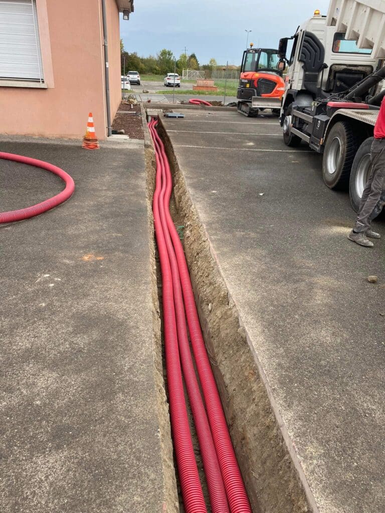 Installation de fourreaux électriques dans une tranchée Pose de gaines rouges annelées dans une tranchée sur un parking, avec pelleteuse et camion en arrière-plan.