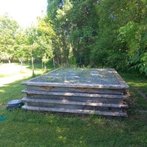 Bac de Compostage en Bois avec Grillage pour Jardin Bac en bois à niveaux avec grillage et herbes folles à côté d'une fosse septique dans un jardin ensoleillé.