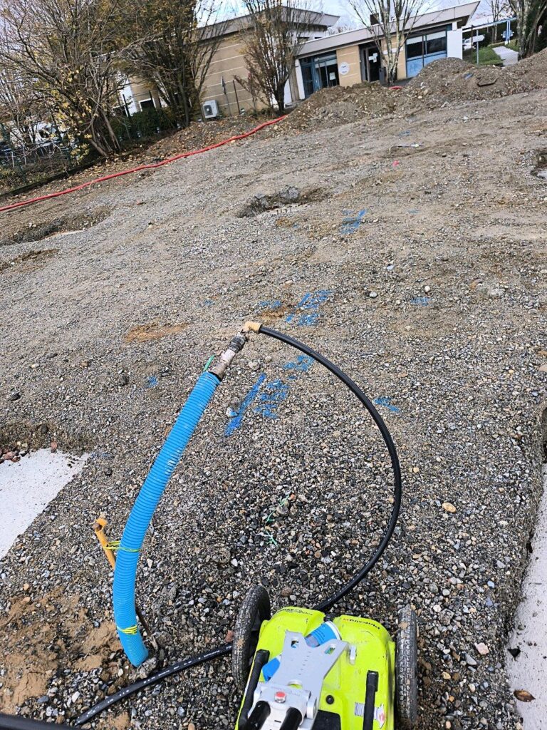 Chantier avec gaine bleue, tuyau noir et pompe d'essai sur gravier. Marquages bleus au sol et bâtiment au fond.